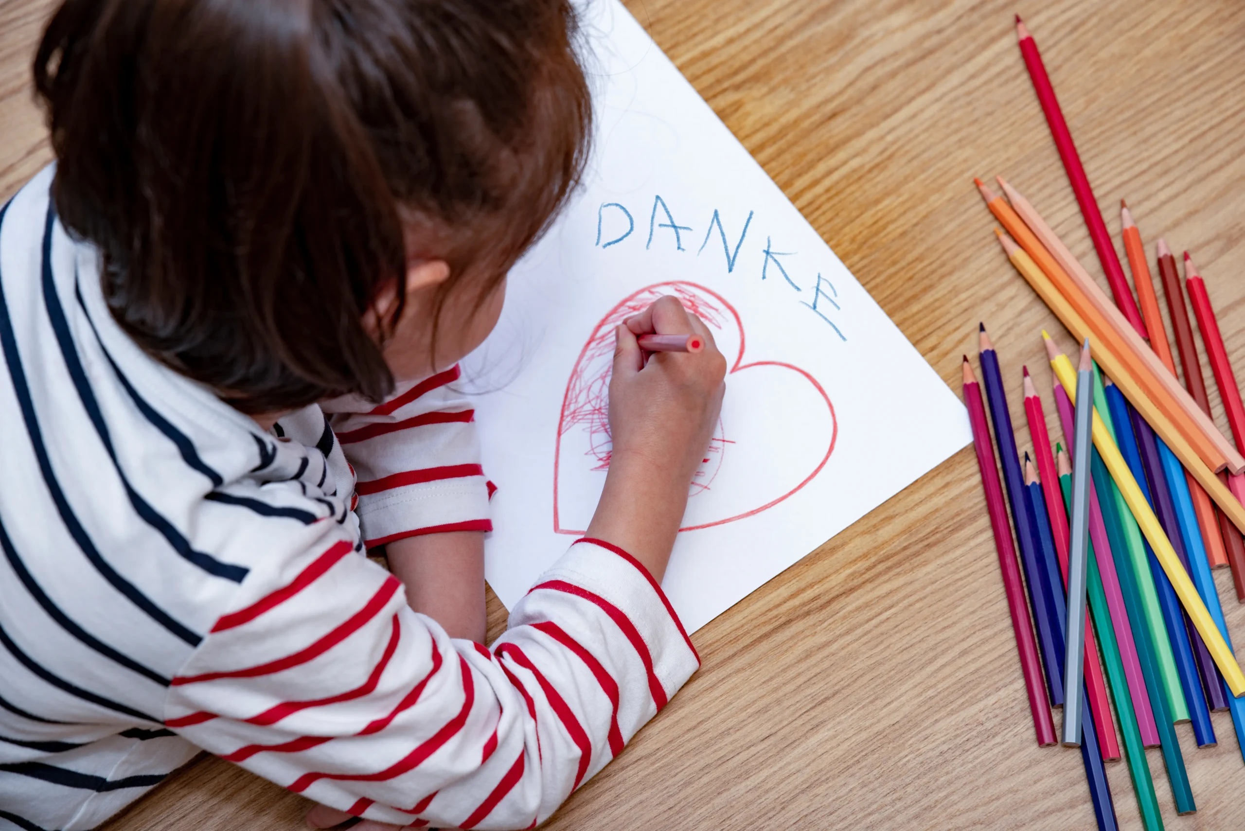 Ein Kind, dass ein Herz auf ein Blatt Papier malt. Über dem Herz steht in Kinderschrift das Wort Danke! Neben dem Blatt liegen verschiedene Buntstifte.